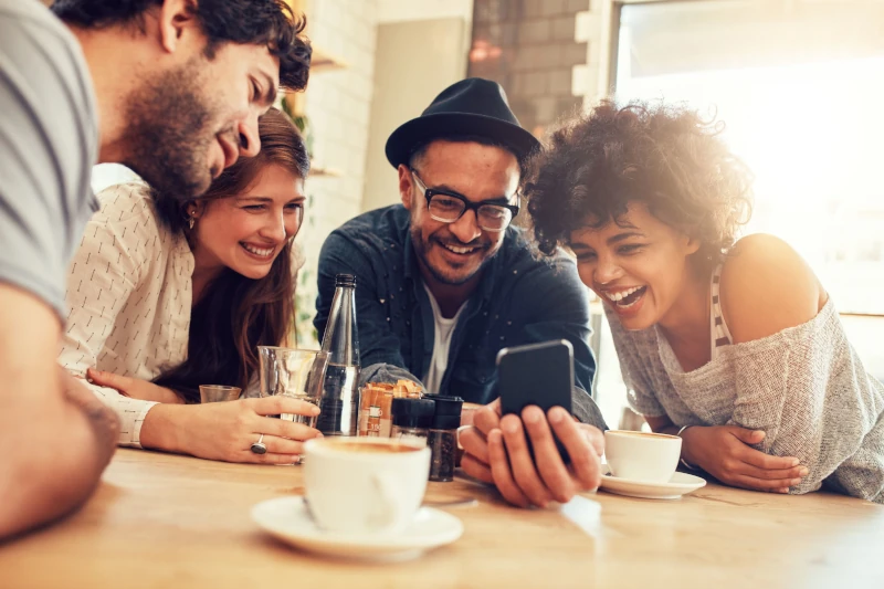 Vier Freunde lachen gemeinsam in einem Café und schauen auf ein Smartphone am Tisch.