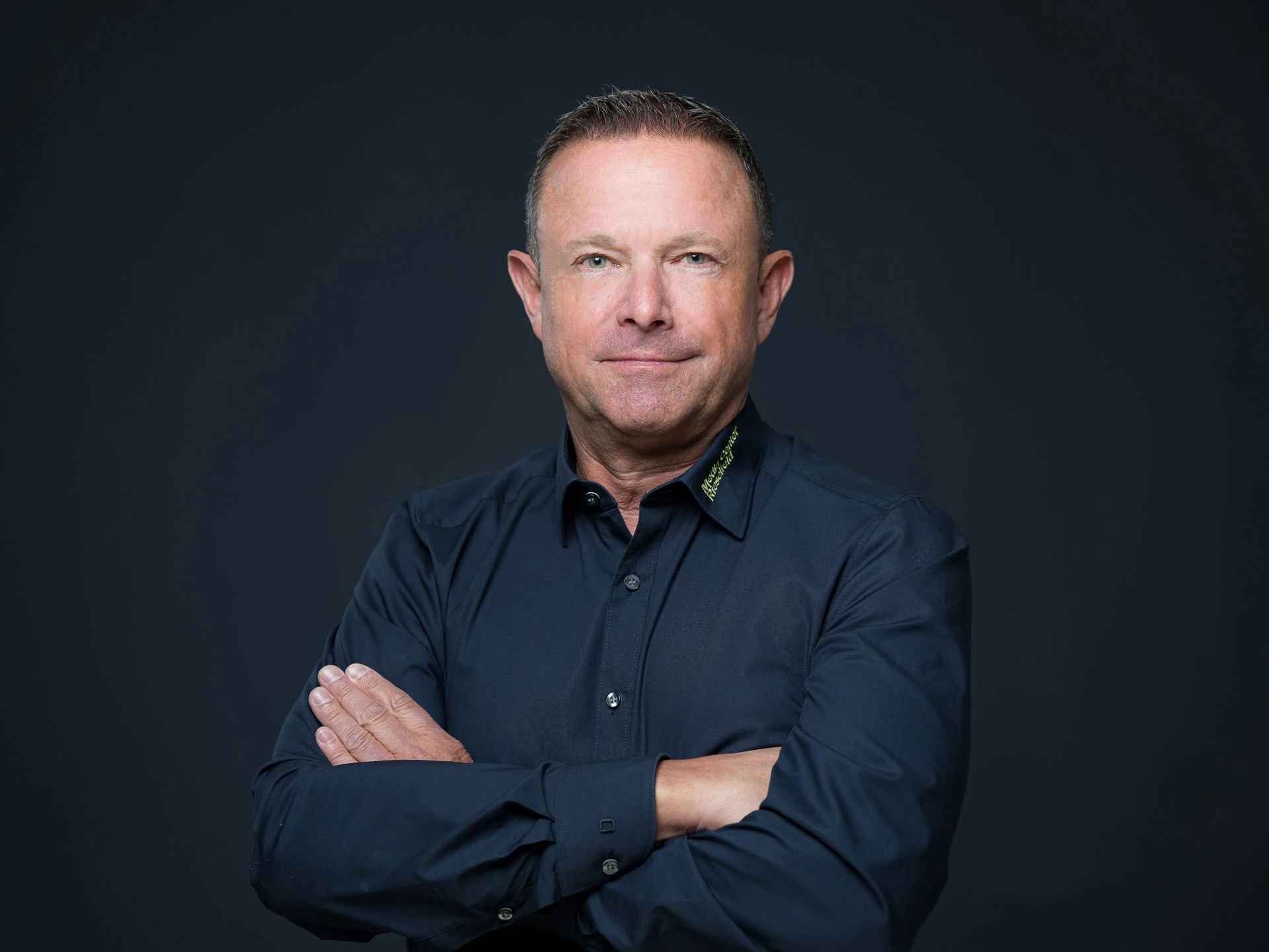 Porträt eines Mannes im dunkelblauen Hemd mit verschränkten Armen vor dunklem Hintergrund.